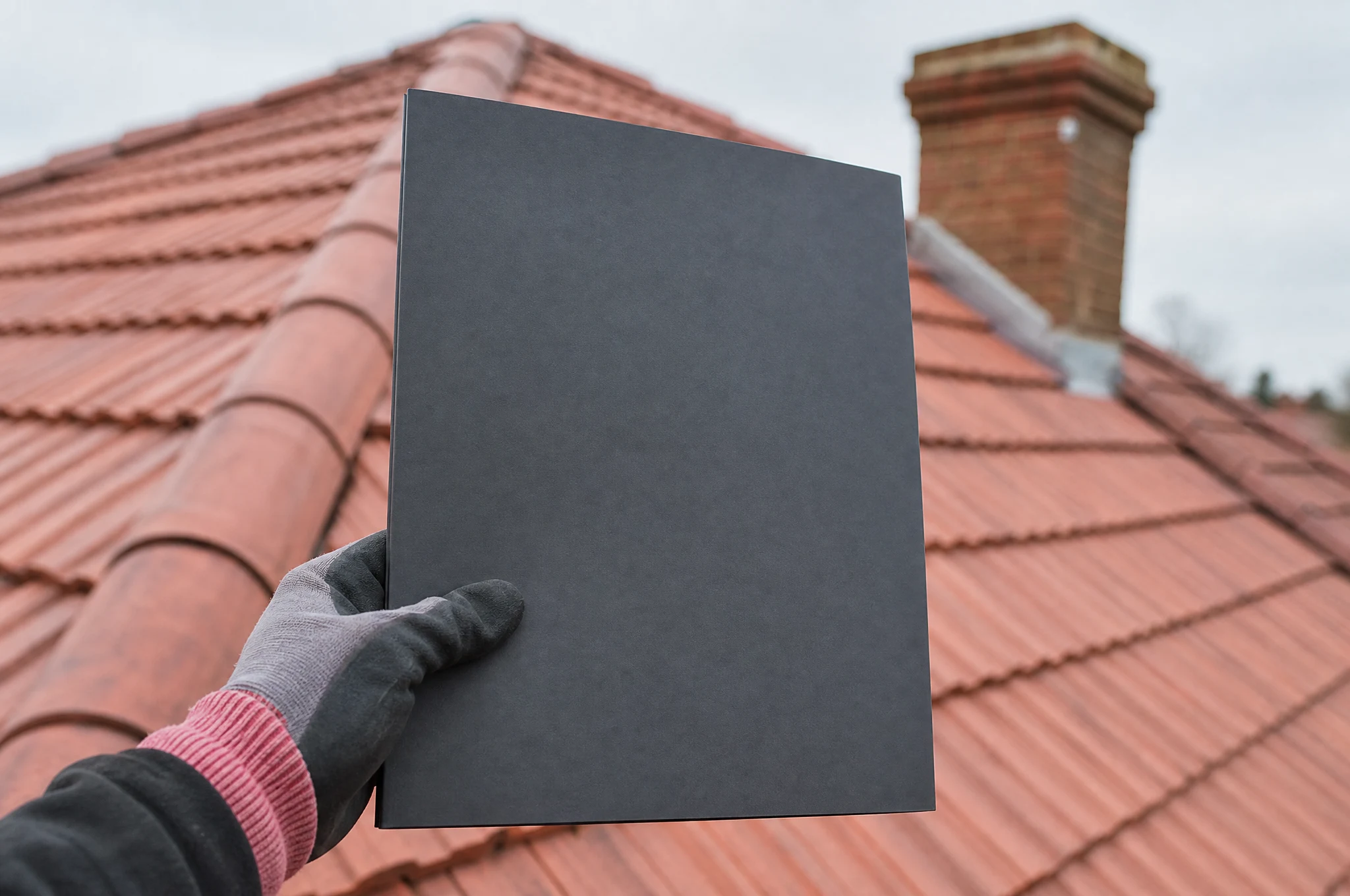 A roofer holding a guarantee document folder in front of a freshly tiled roof and brick chimney