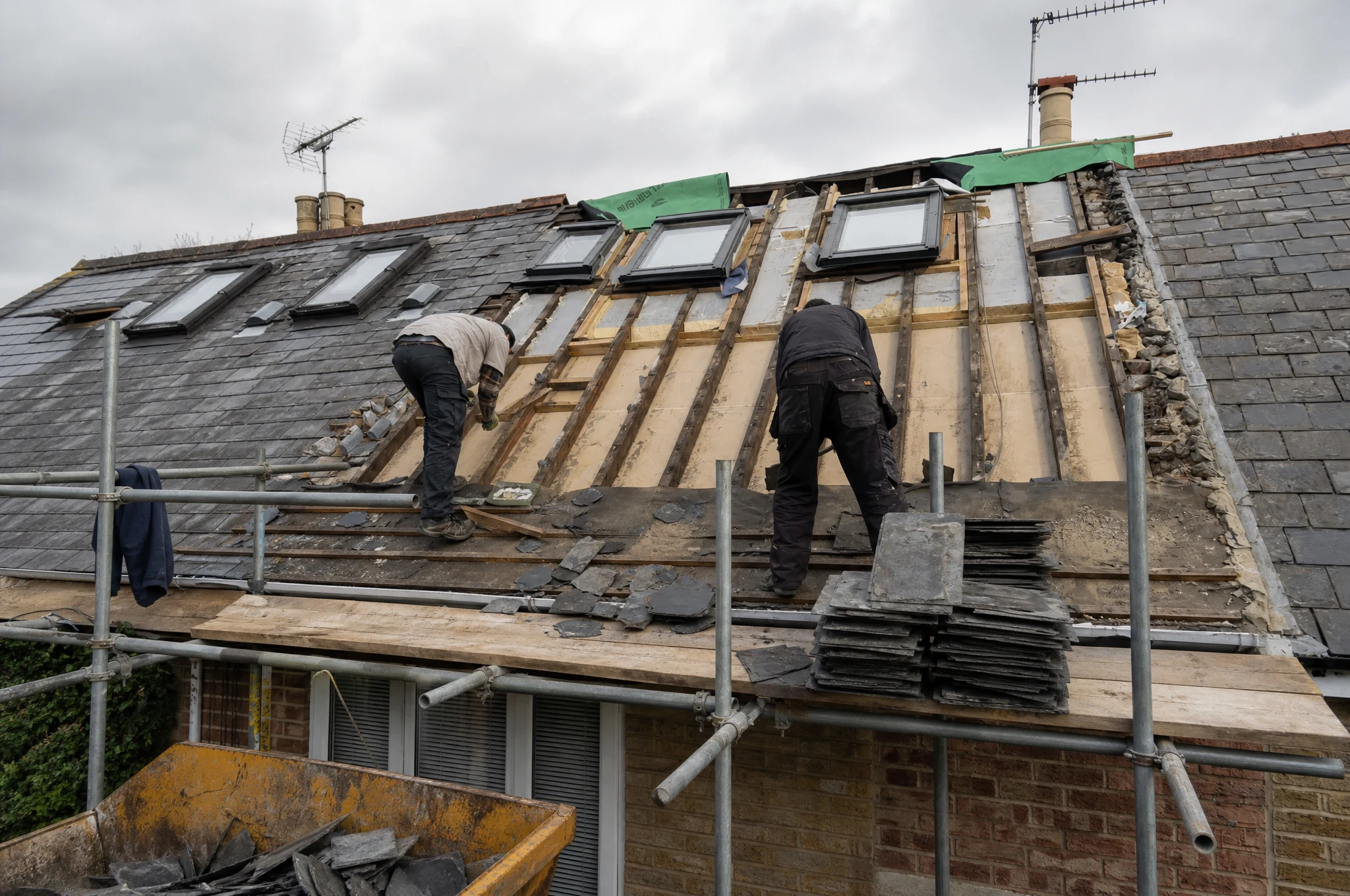 Roofers stripping old slate tiles from a UK roof during the first stage of a re-roof, scaffolding and skip visible below